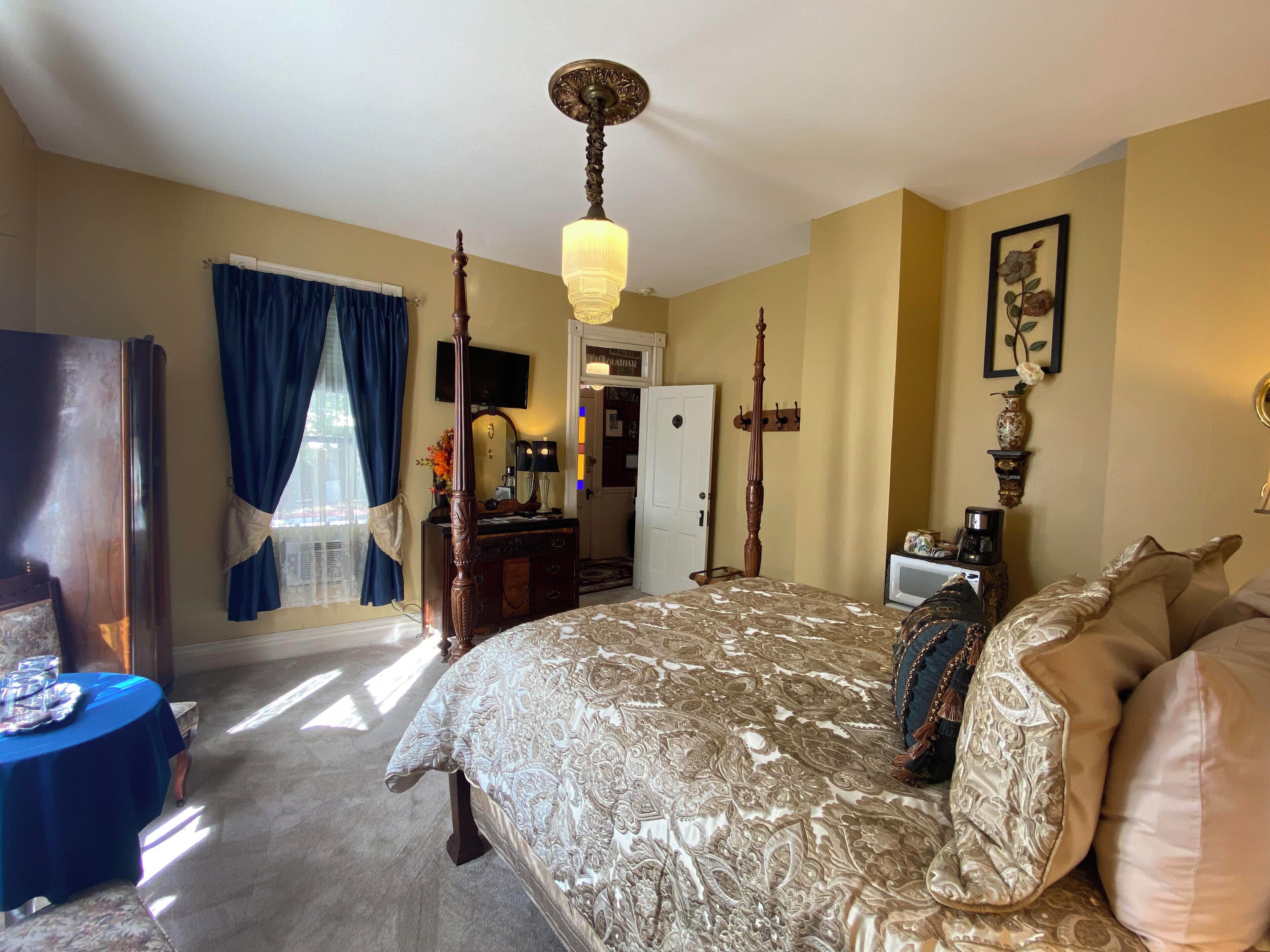 A bedroom with a large bed, a wooden wardrobe, a desk with a chair, a television, and a door leading to another room.