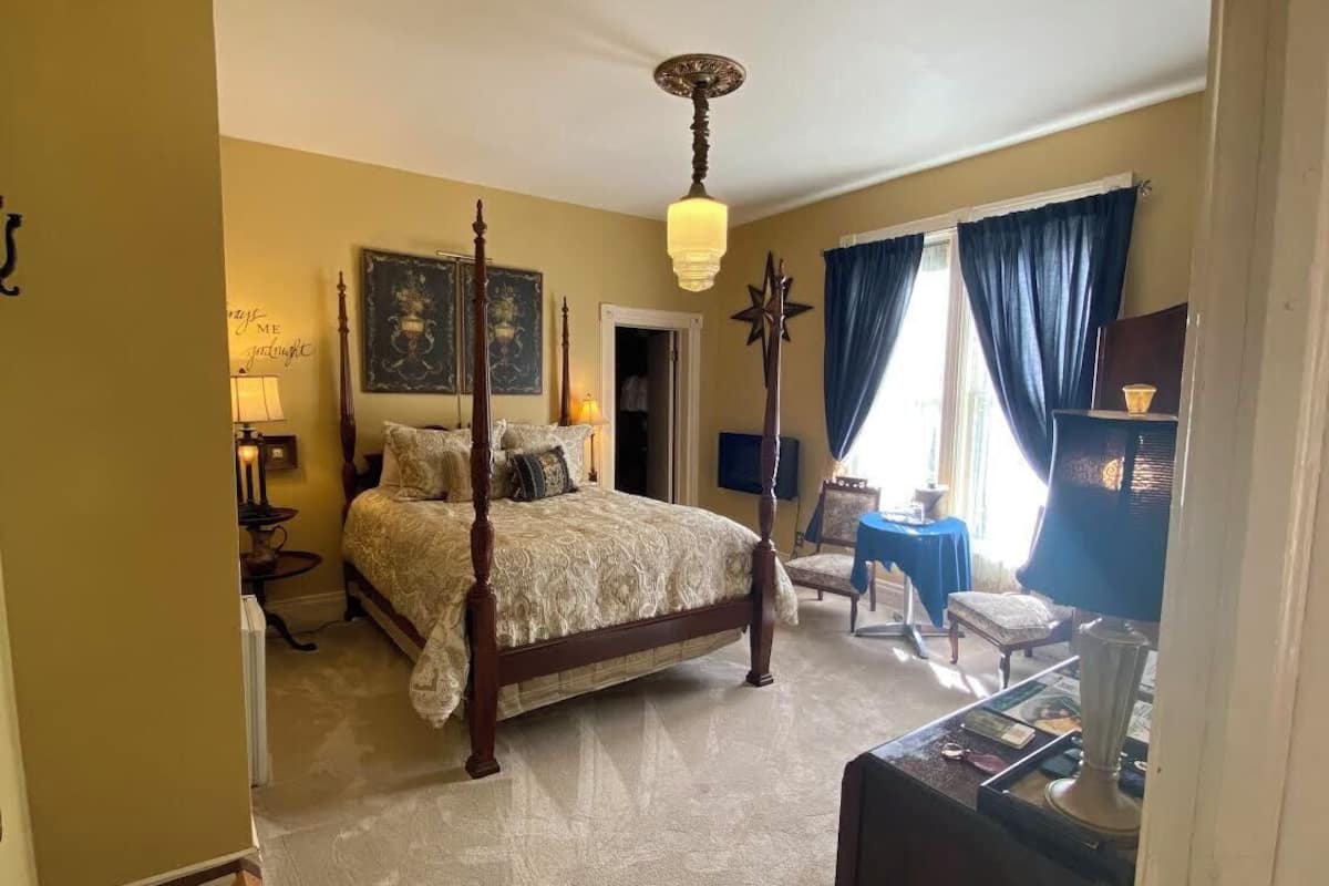 A bedroom with a four-poster bed, a chandelier, a desk with a lamp, and a view of the outside through a window with curtains.