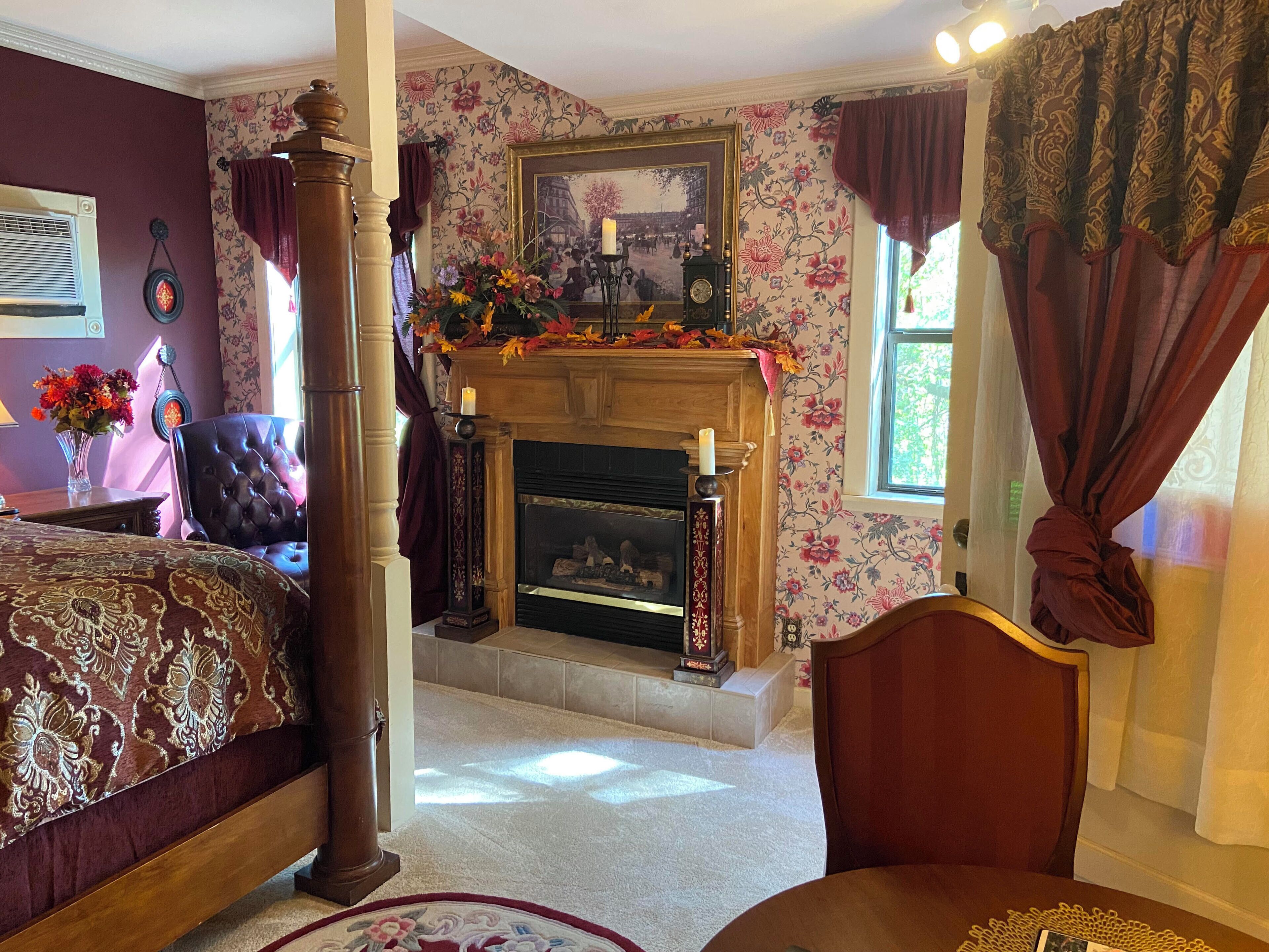 A room with a fireplace, floral wallpaper, a bed, a chair, and a table.