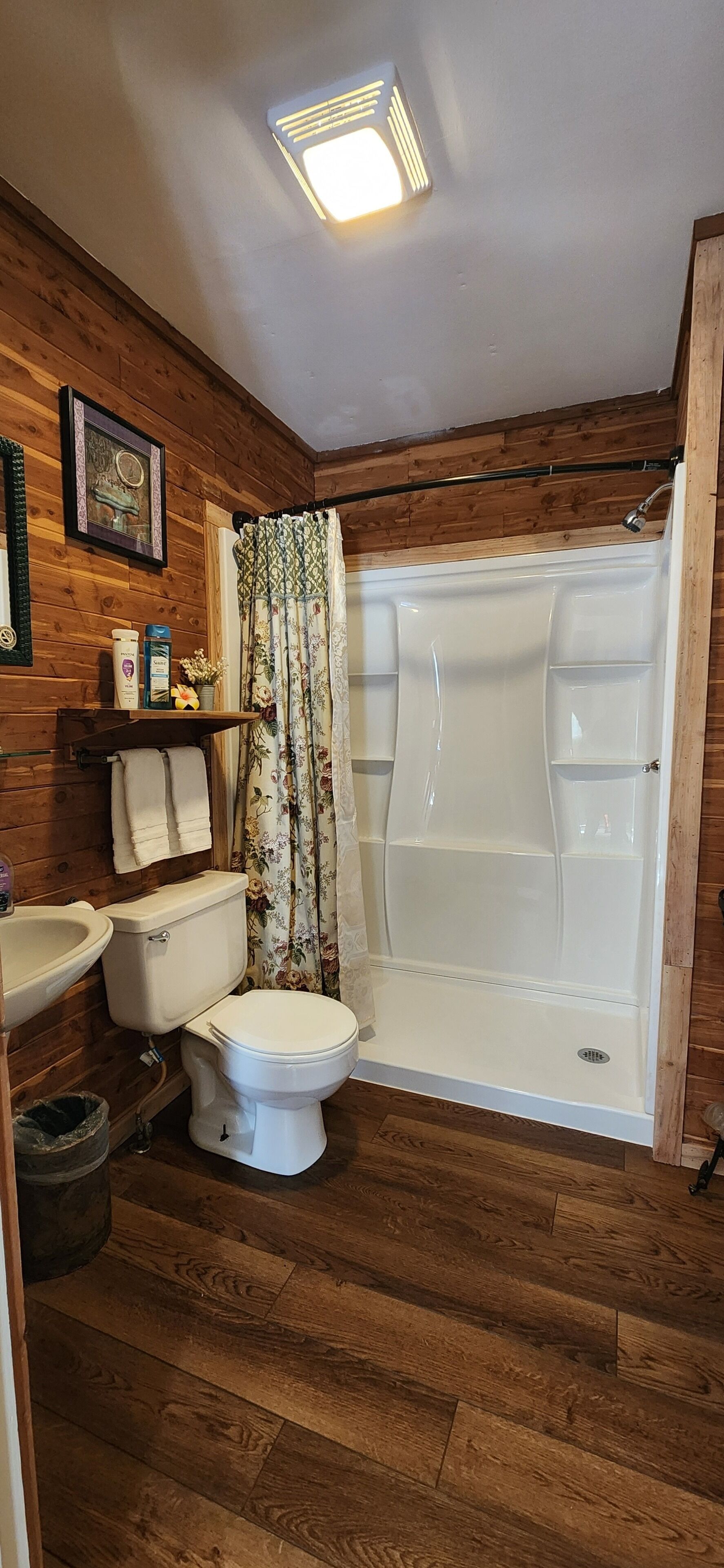 A bathroom with wooden walls, a toilet, a sink, a shower with a curtain, and a small shelf with toiletries.
