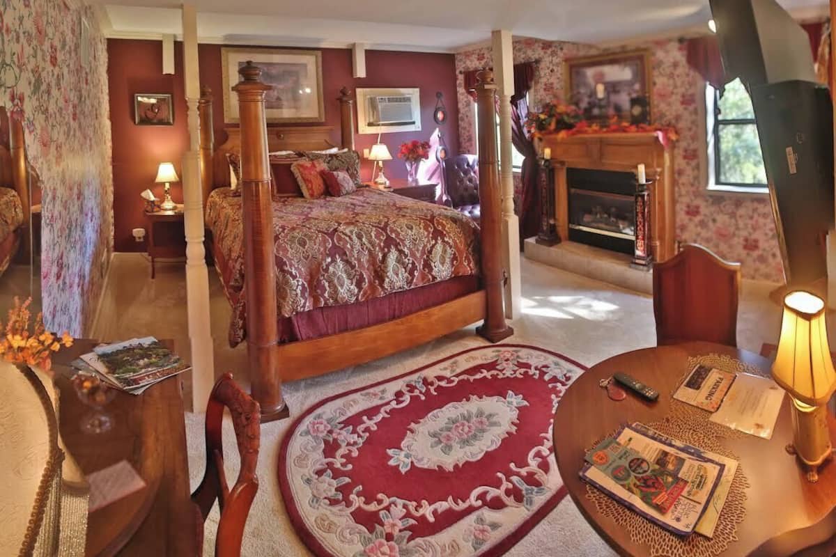 A traditional bedroom with a four-poster bed, a fireplace, and a round table with a lamp.