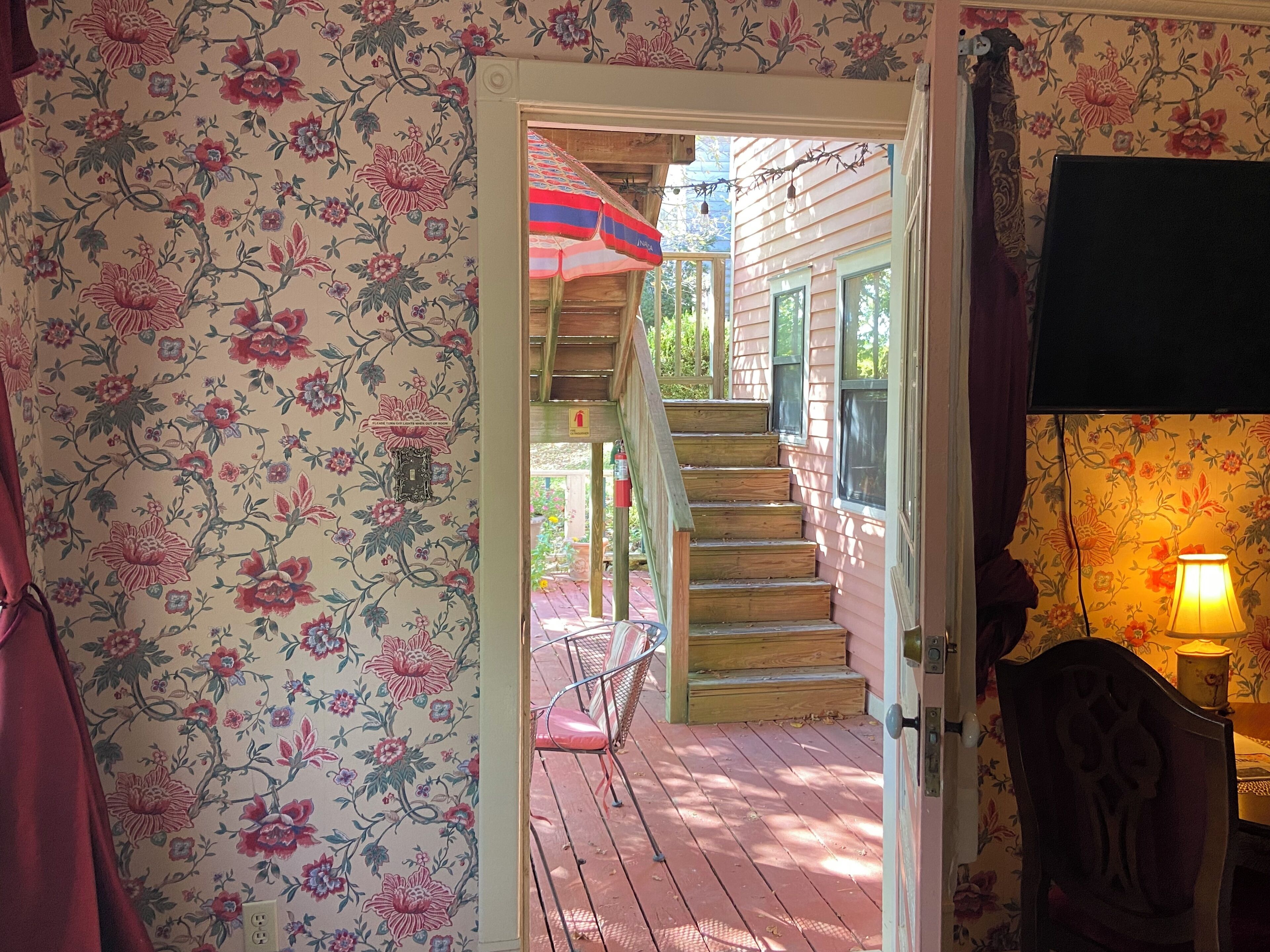 Una escalera de madera conduce a un porche con una pared cubierta de papel tapiz floral, un paraguas rojo y una lámpara a la derecha.