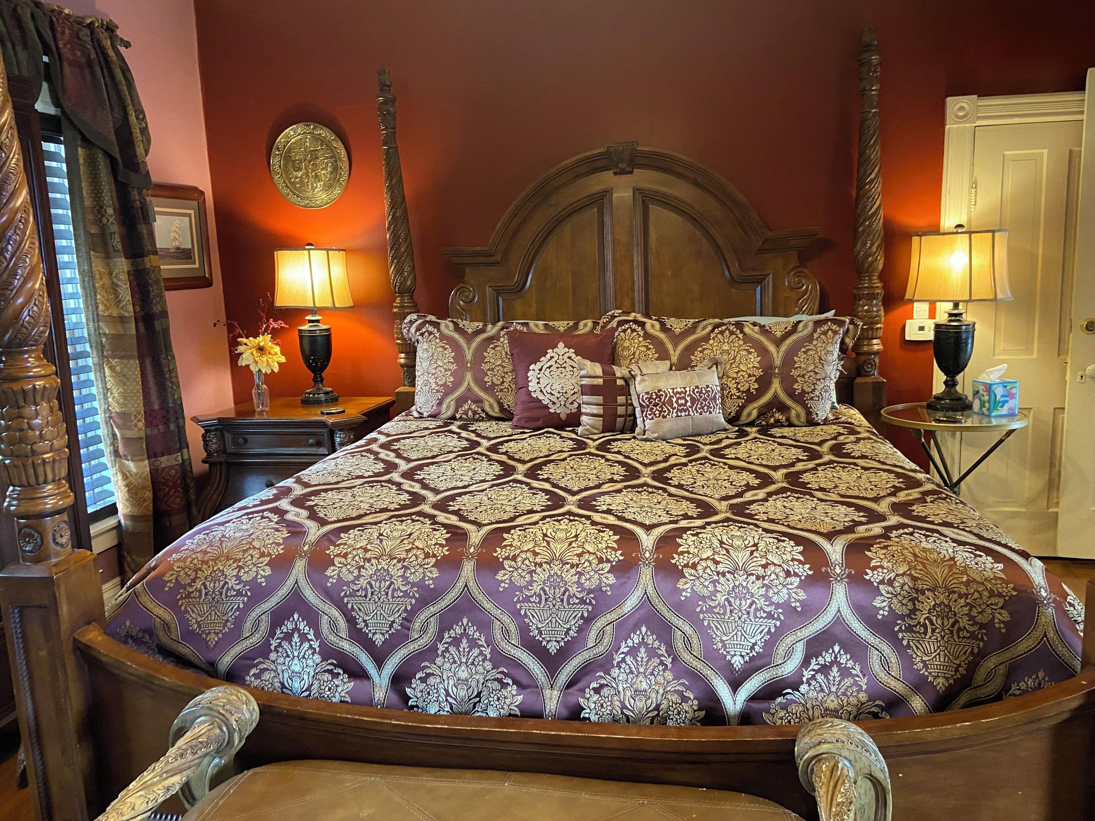 A large bed with a patterned bedspread, two bedside tables with lamps, a wooden headboard, and a curtain on the left.