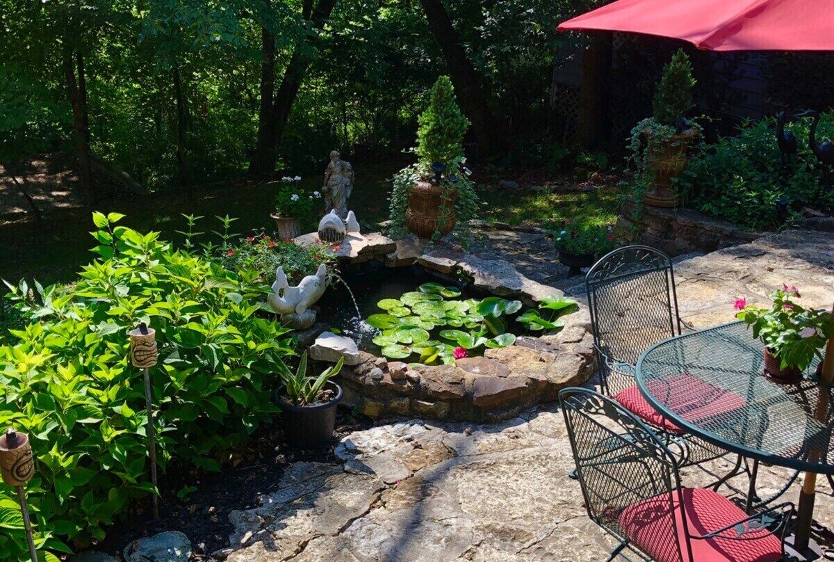 Un patio jardín con una fuente de piedra, plantas en macetas y una mesa metálica con cojines rojos.