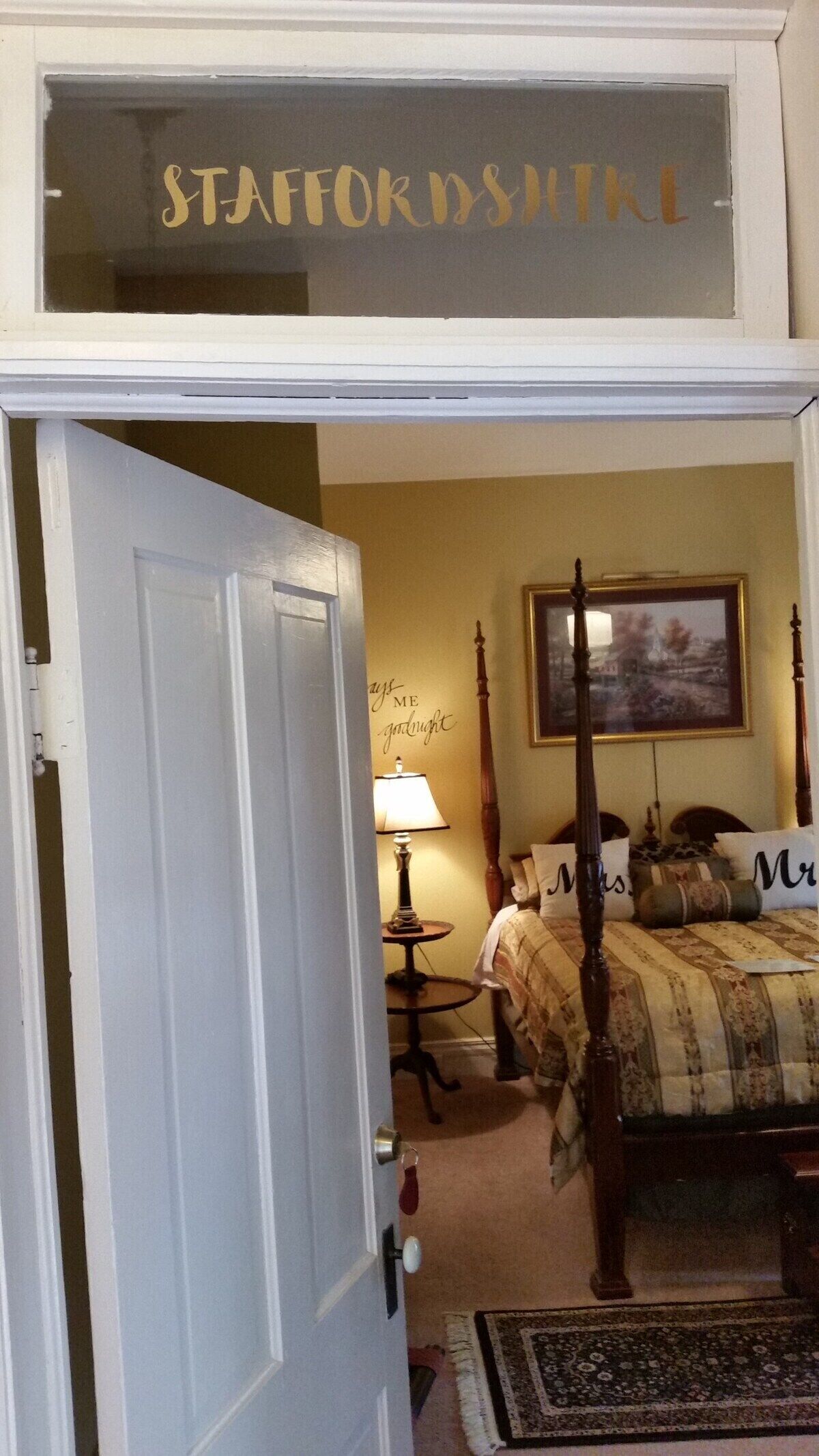 A bedroom with a bed, a bedside table with a lamp, a wall clock, and a framed picture.