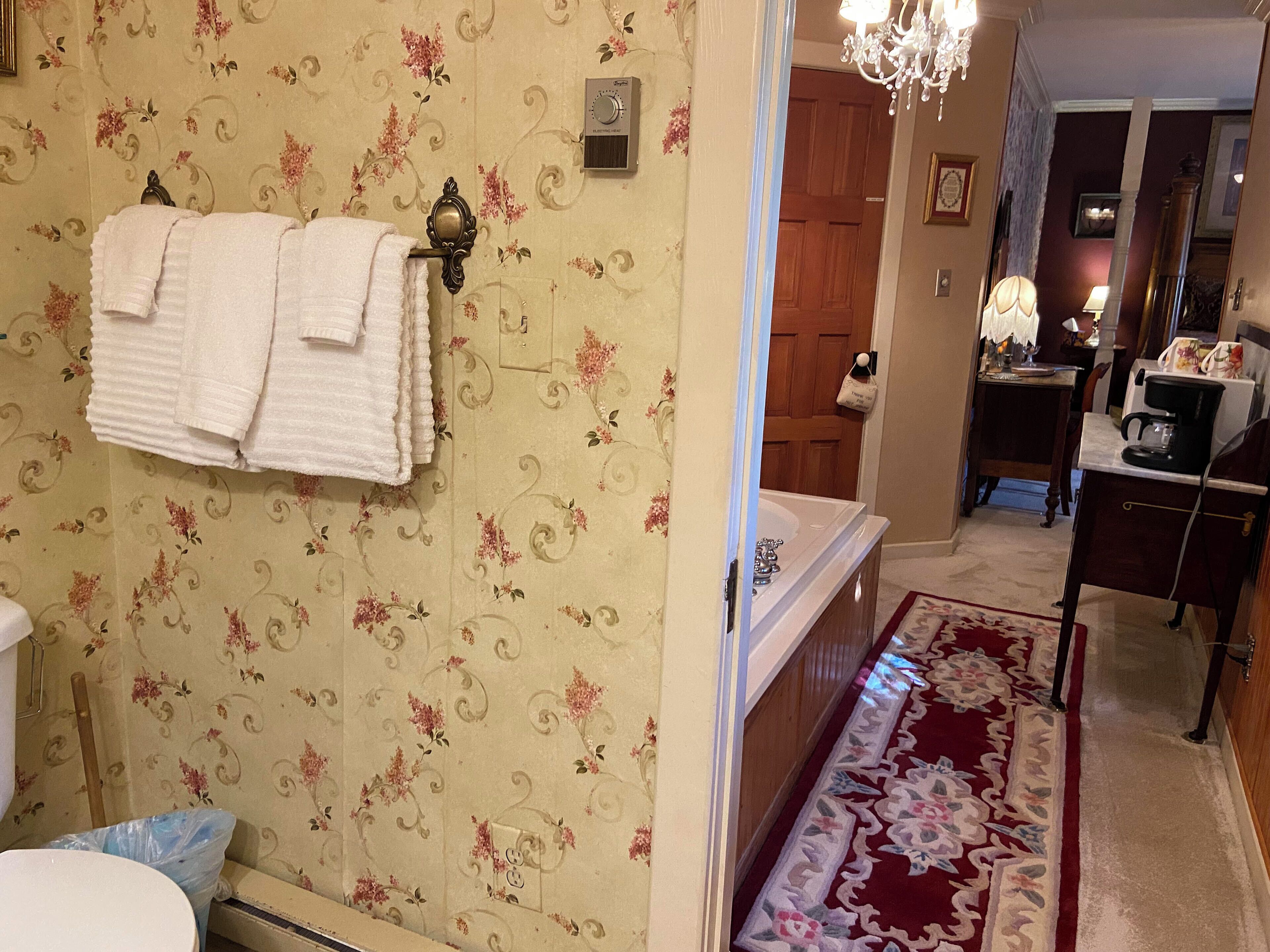A bathroom with floral wallpaper, a white bathtub, and a red patterned rug leading to a hallway.