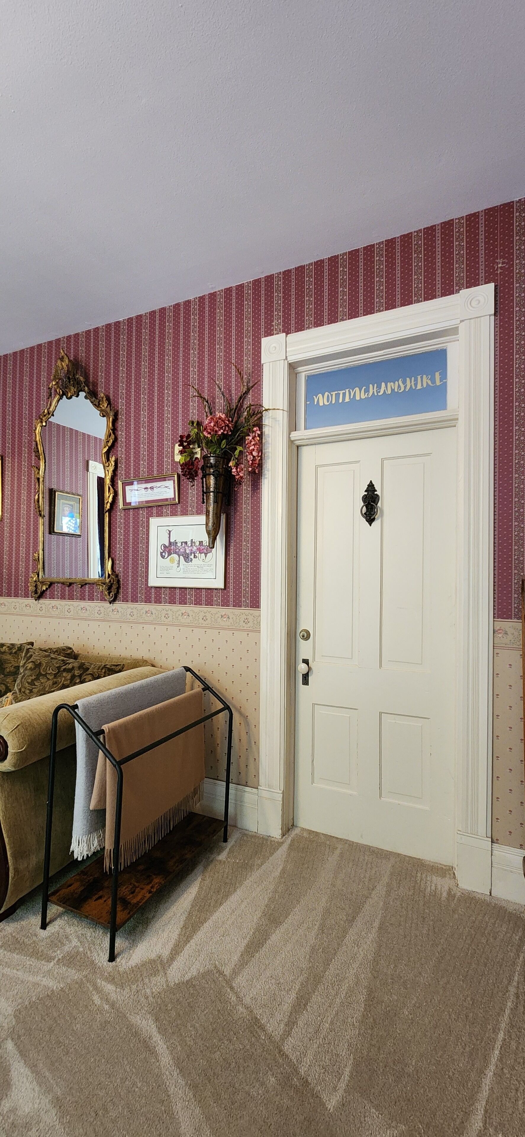 Quarto com papel de parede estampado, porta branca com a placa 'Nottinghamshire', espelho, quadros emoldurados e um banco com mantas.