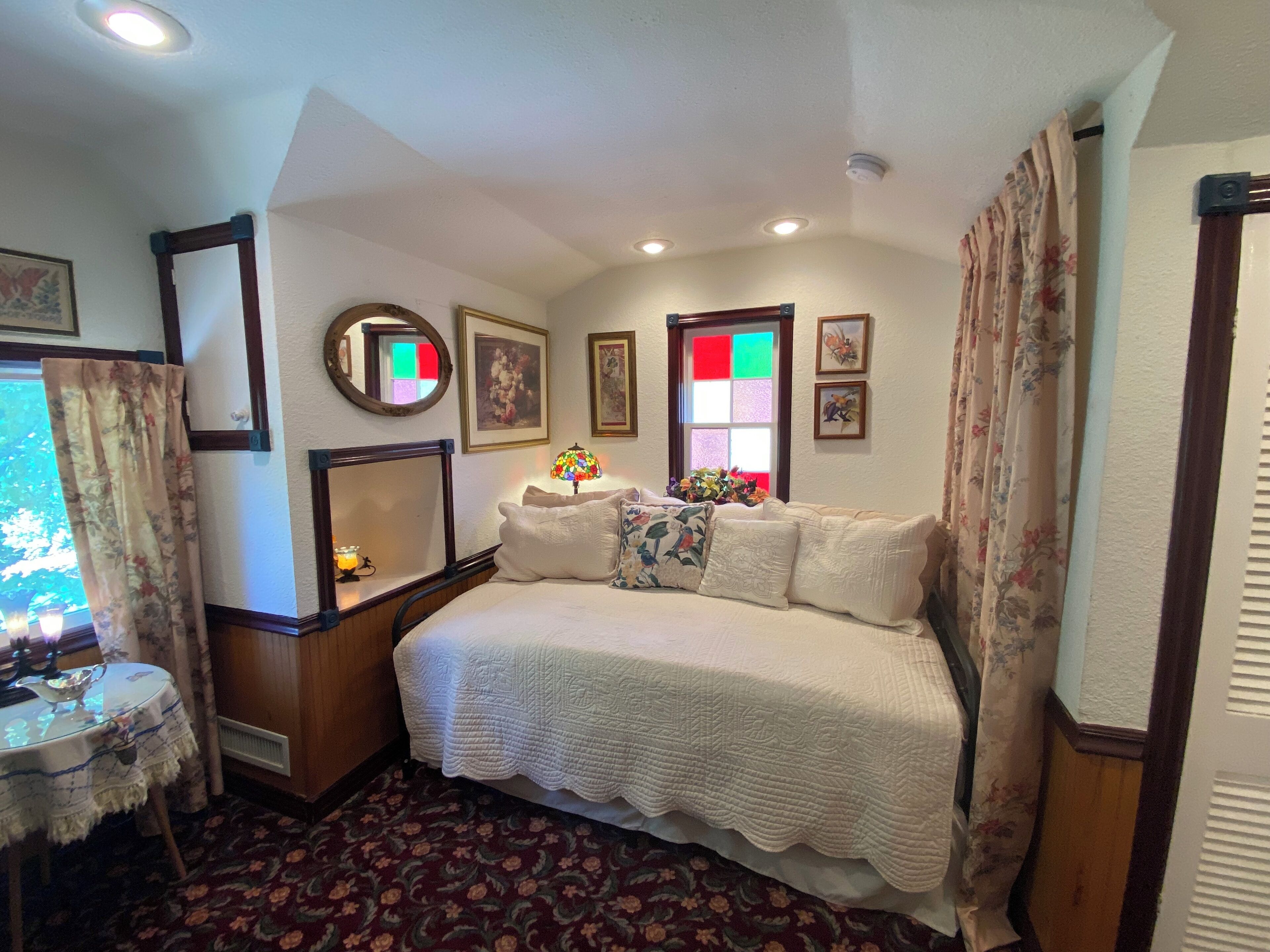 Un dormitorio con una cama, una mesita de noche, un espejo, una ventana con vistas y una cortina de estampado floral.