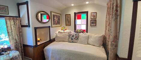 Un dormitorio con una cama, una mesita de noche, un espejo, una ventana con vistas y una cortina de estampado floral.