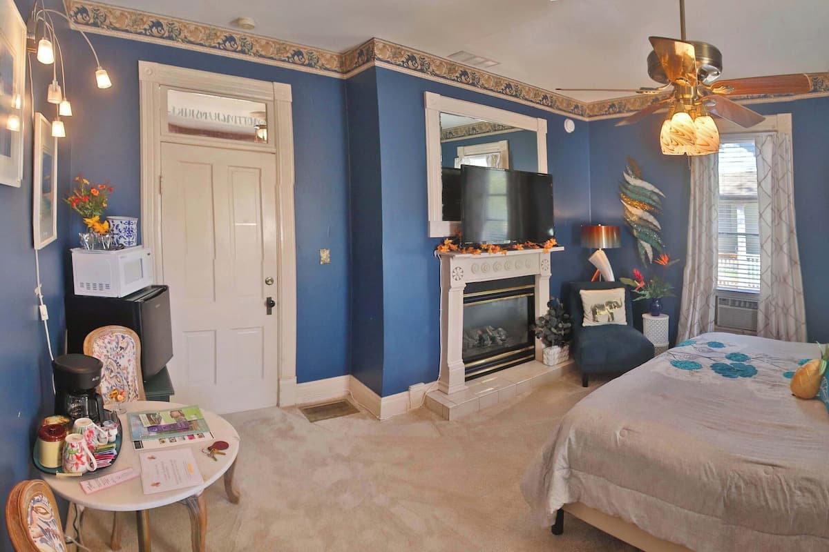 A bedroom with a fireplace, a bed with multiple pillows, a television, and a ceiling fan.