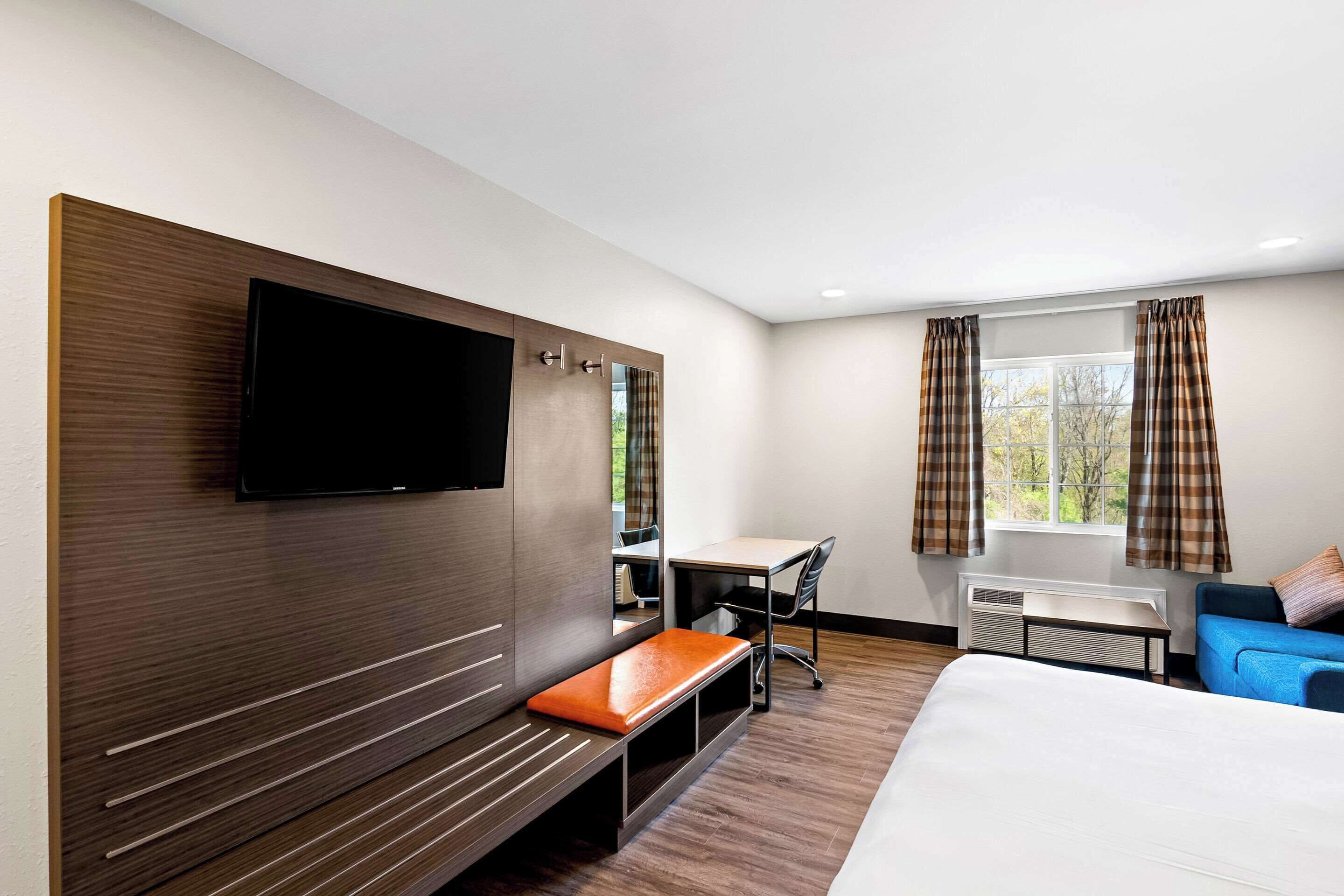 A modern hotel room with a large bed, a flat-screen TV mounted on a wooden panel, a desk with a chair, a bench, and a window with curtains.