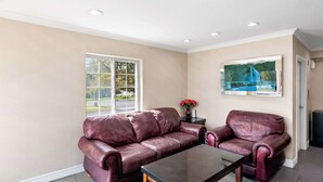 A living room with a beige wall, a glass coffee table, two leather sofas, a small table with a vase of flowers, and a painting of a blue sky and trees. - Econo Lodge Bethel - Danbury (Bethel)