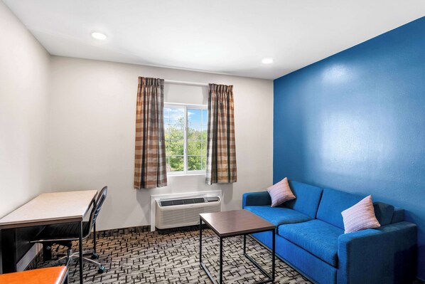 A room with a blue sofa, a small table, a desk with a chair, a window with curtains, and a patterned floor.