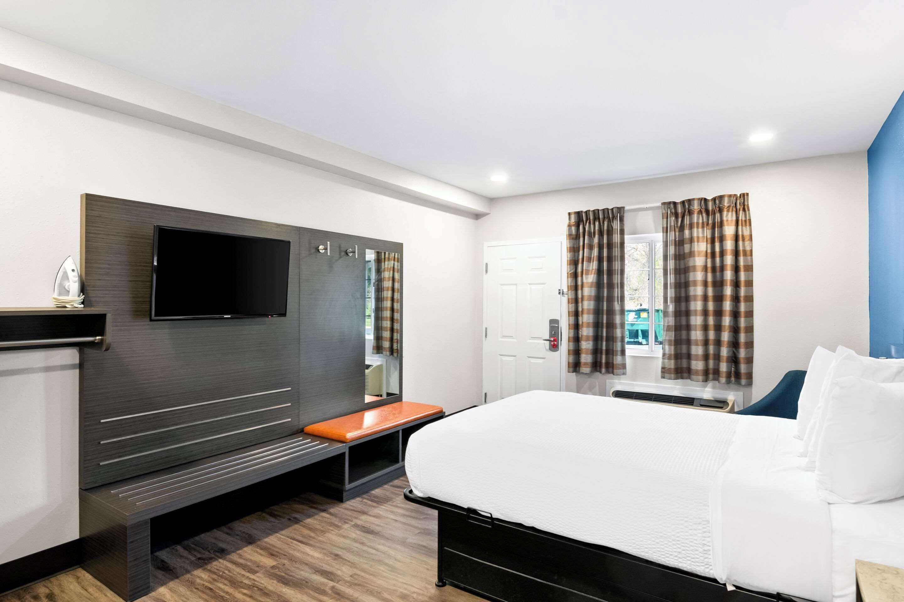 A hotel room with a bed, a TV mounted on the wall, a bench, and a window with curtains.