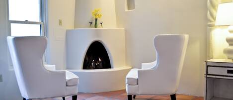 A room with two white armchairs, a fireplace, a window, and a lamp.