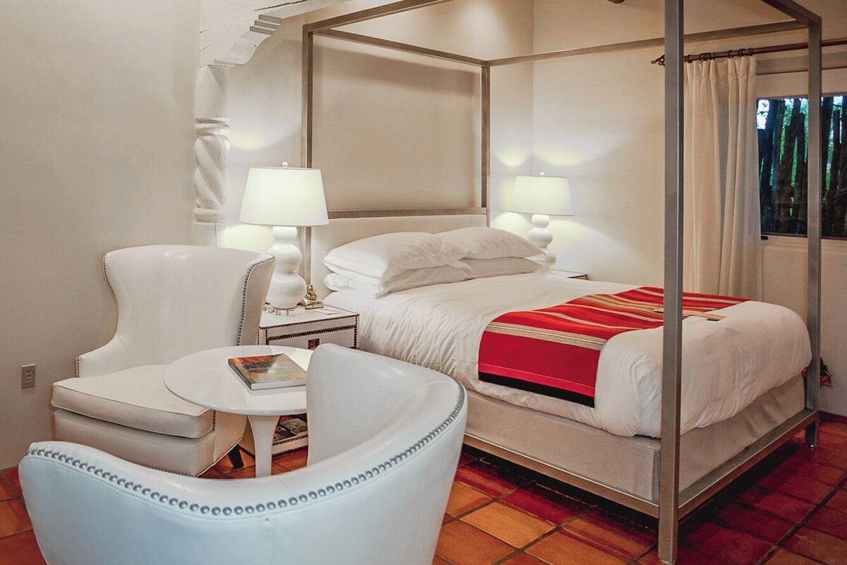A four-poster bed with white linens and a red throw, a white armchair, and a small table with a lamp.