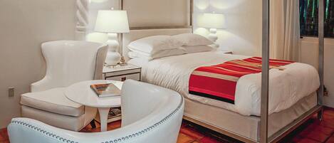 Una cama con dosel con sábanas blancas y una manta roja, un sillón blanco y una mesita de noche con una lámpara.