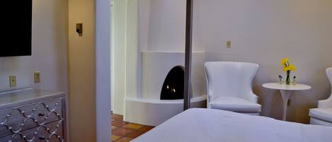 Una habitación de hotel con una cama, un sillón blanco, una mesita redonda con un florero y una chimenea, además de un televisor montado en la pared.