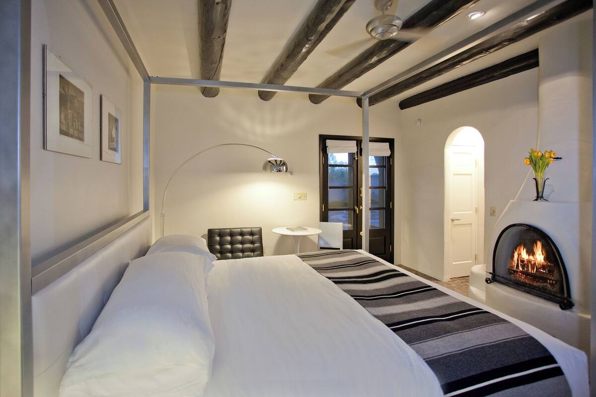 A hotel room with a large bed, a small table, a chair, a fireplace, and framed pictures on the wall.