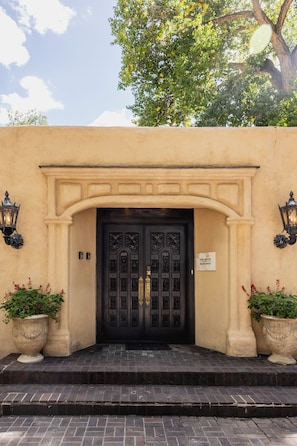 A building entrance with a black ornate door, two wall-mounted lanterns, and potted plants on either side.