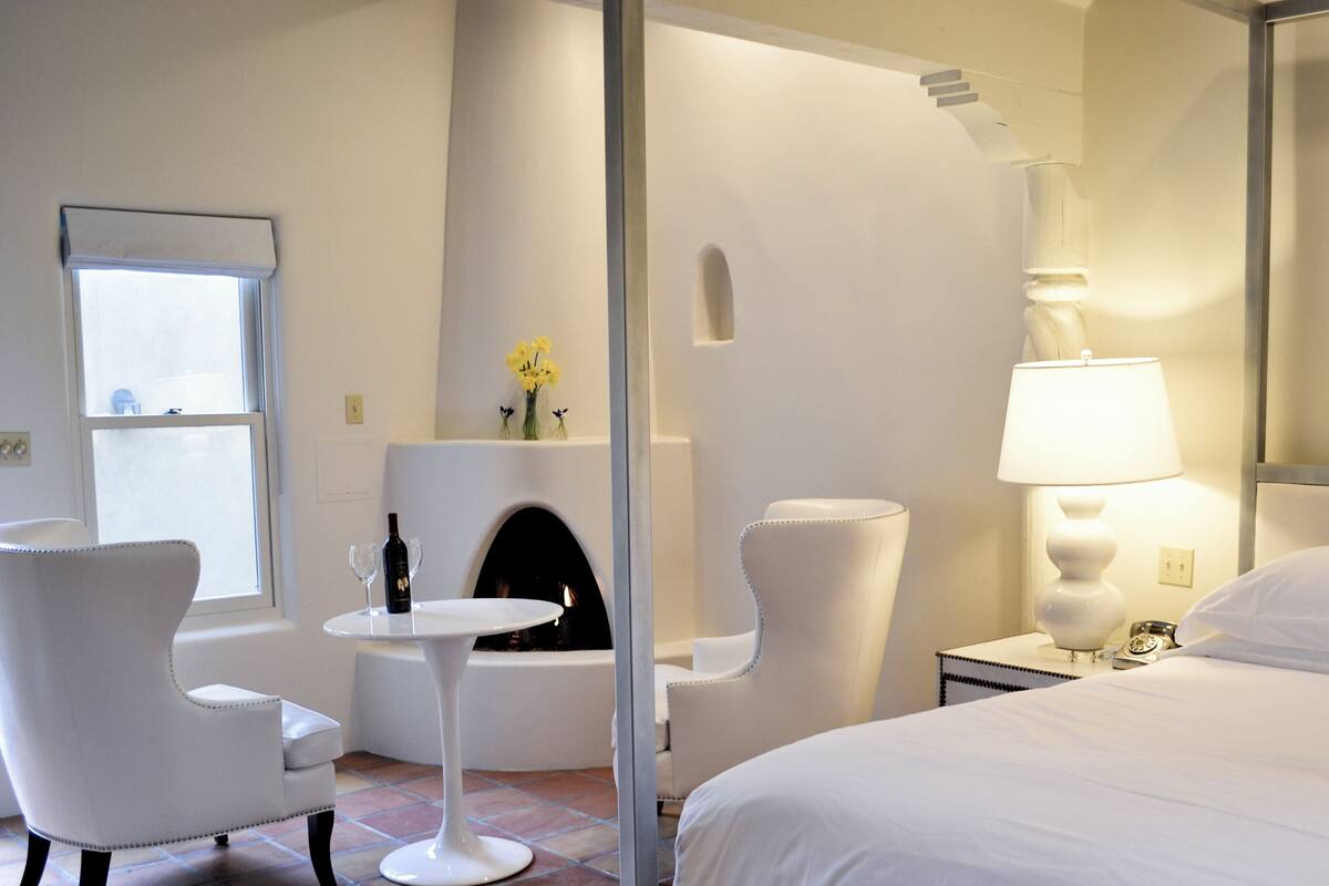 A hotel room with a bed, a white chair, a small table, a lamp, and a fireplace.