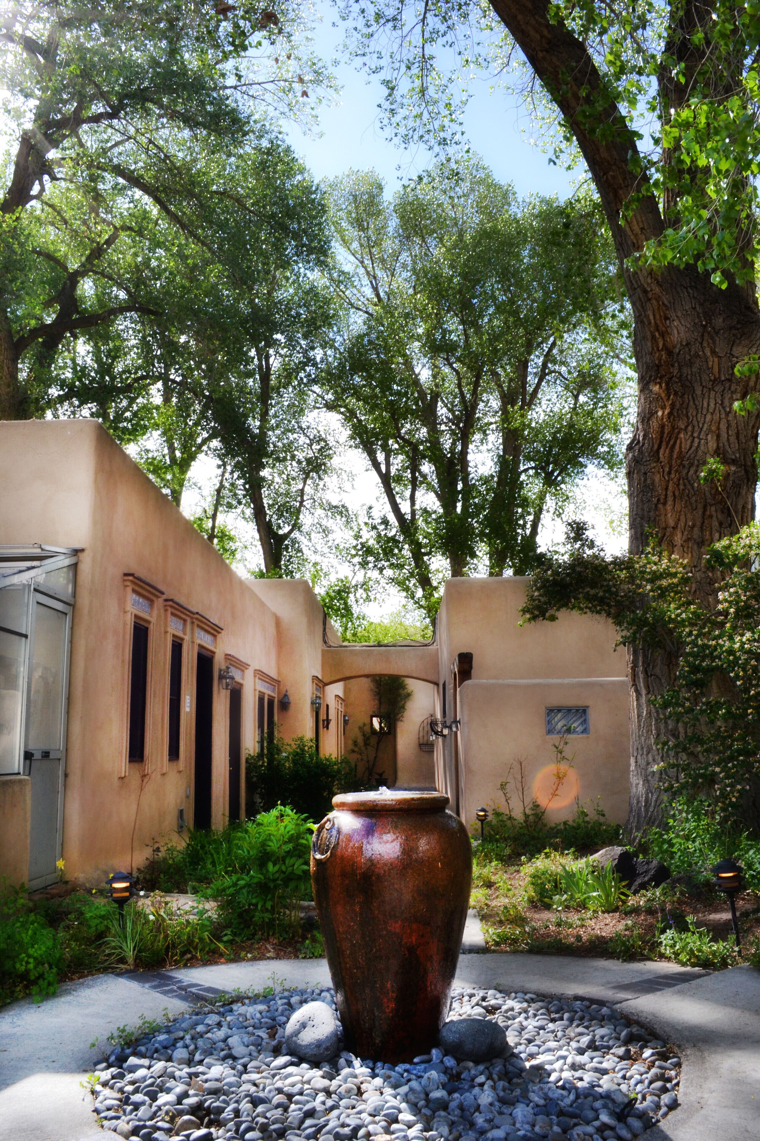 A rustic outdoor area with a large copper pot, surrounded by pebbles, in front of a building with a beige facade and multiple windows.