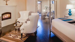 A modern hotel room with a fireplace, a white armchair, a bed, and a view of the hallway.