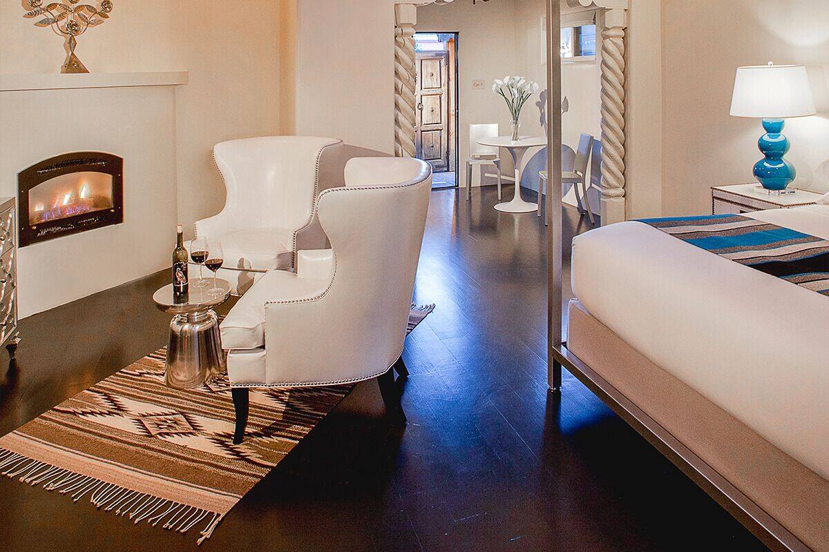 A modern hotel room with a fireplace, a white armchair, a bed, and a view of the hallway.