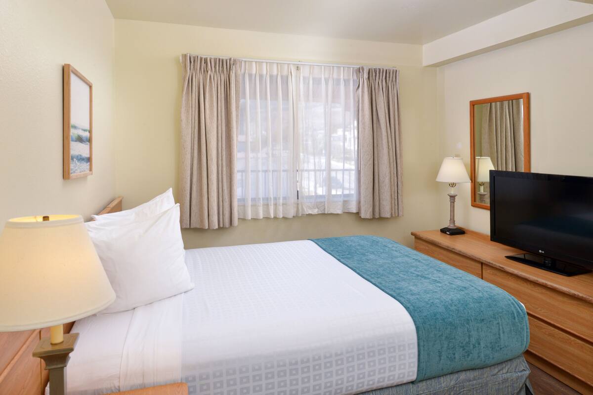 A hotel room with a bed, a television, a desk with a lamp, and a window with curtains.