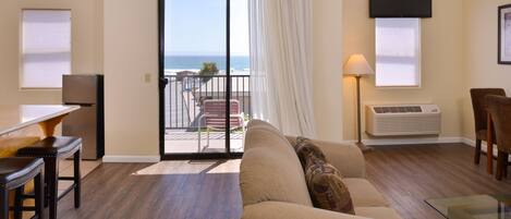 A living room with a sofa, a glass coffee table, a TV, and a balcony with a view of the ocean.