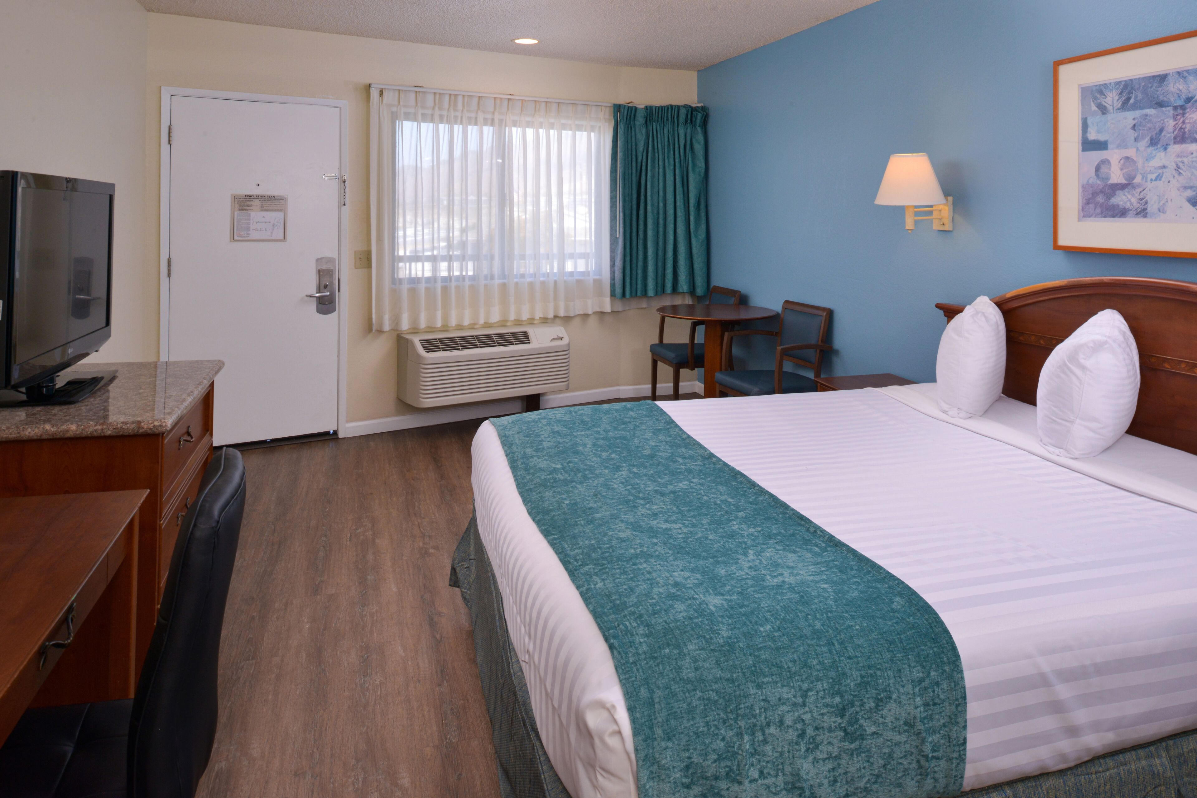 A hotel room with a bed, a desk with a television, a chair, a table, a lamp, and a framed picture on the wall.