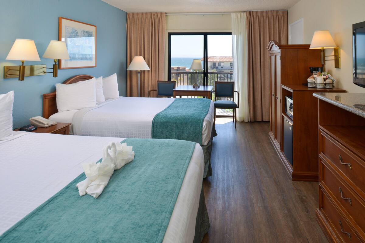A hotel room with two beds, a desk, chairs, a TV, and a view of the ocean.