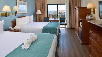 A hotel room with two beds, a desk, chairs, a TV, and a view of the ocean.