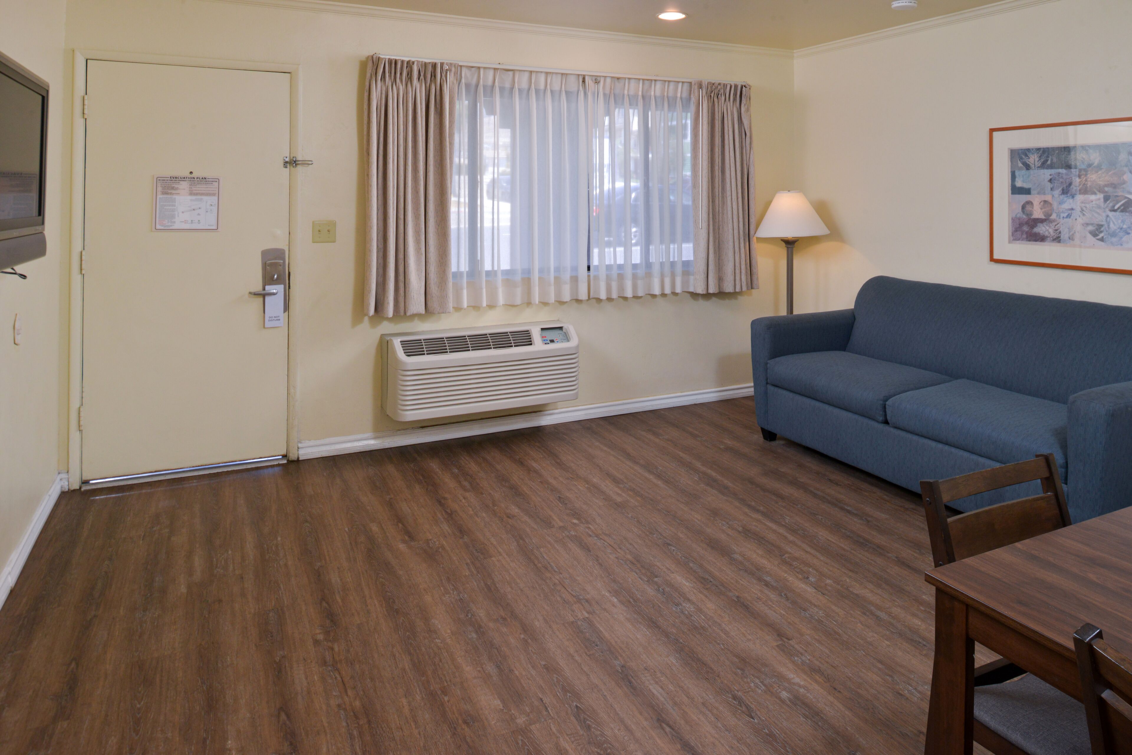 A room with a blue sofa, wooden flooring, a wall-mounted air conditioner, a door with a lock, and a framed picture on the wall.
