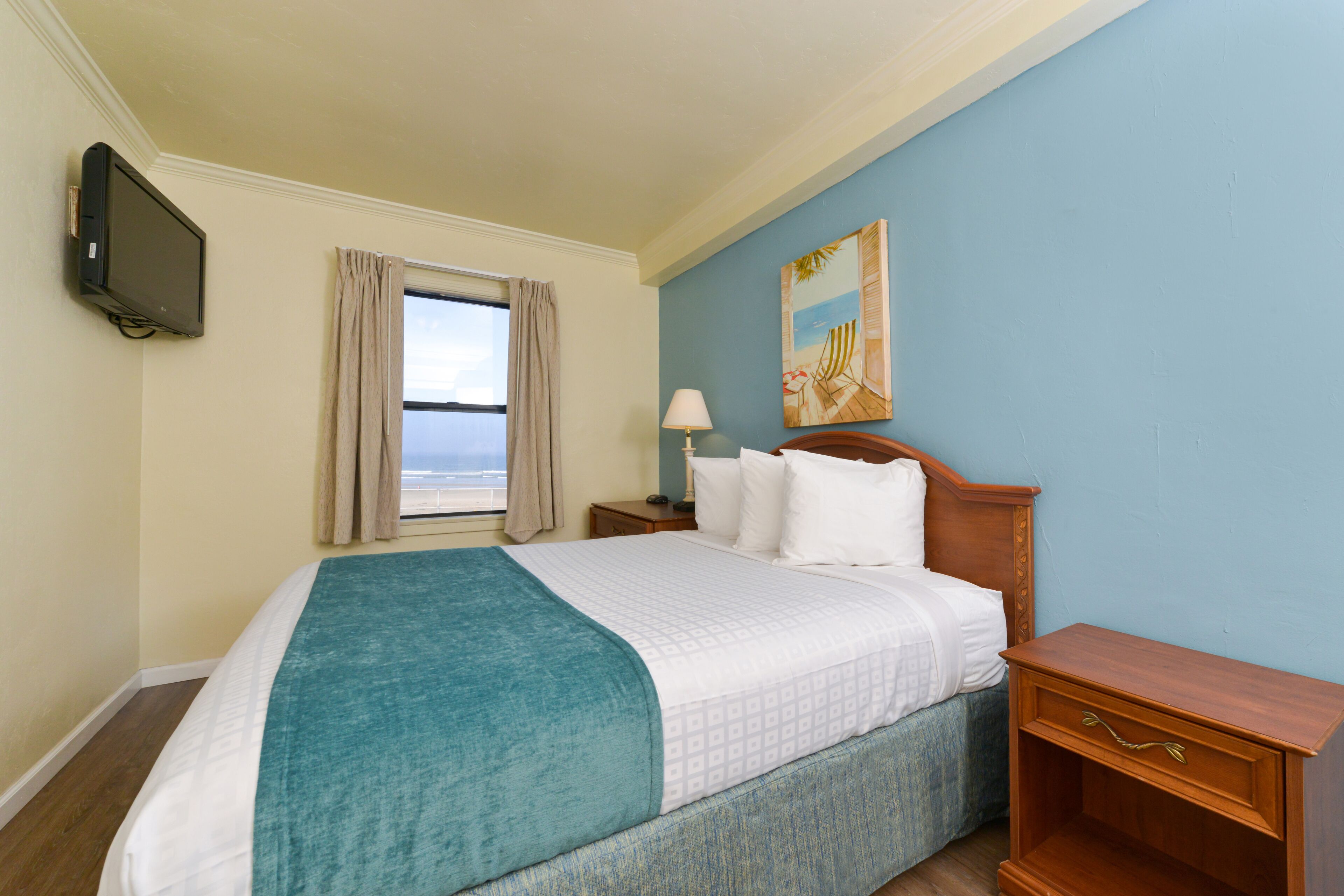 A hotel room with a bed, a TV mounted on the wall, a window with curtains, a nightstand with a lamp, and a painting on the wall.