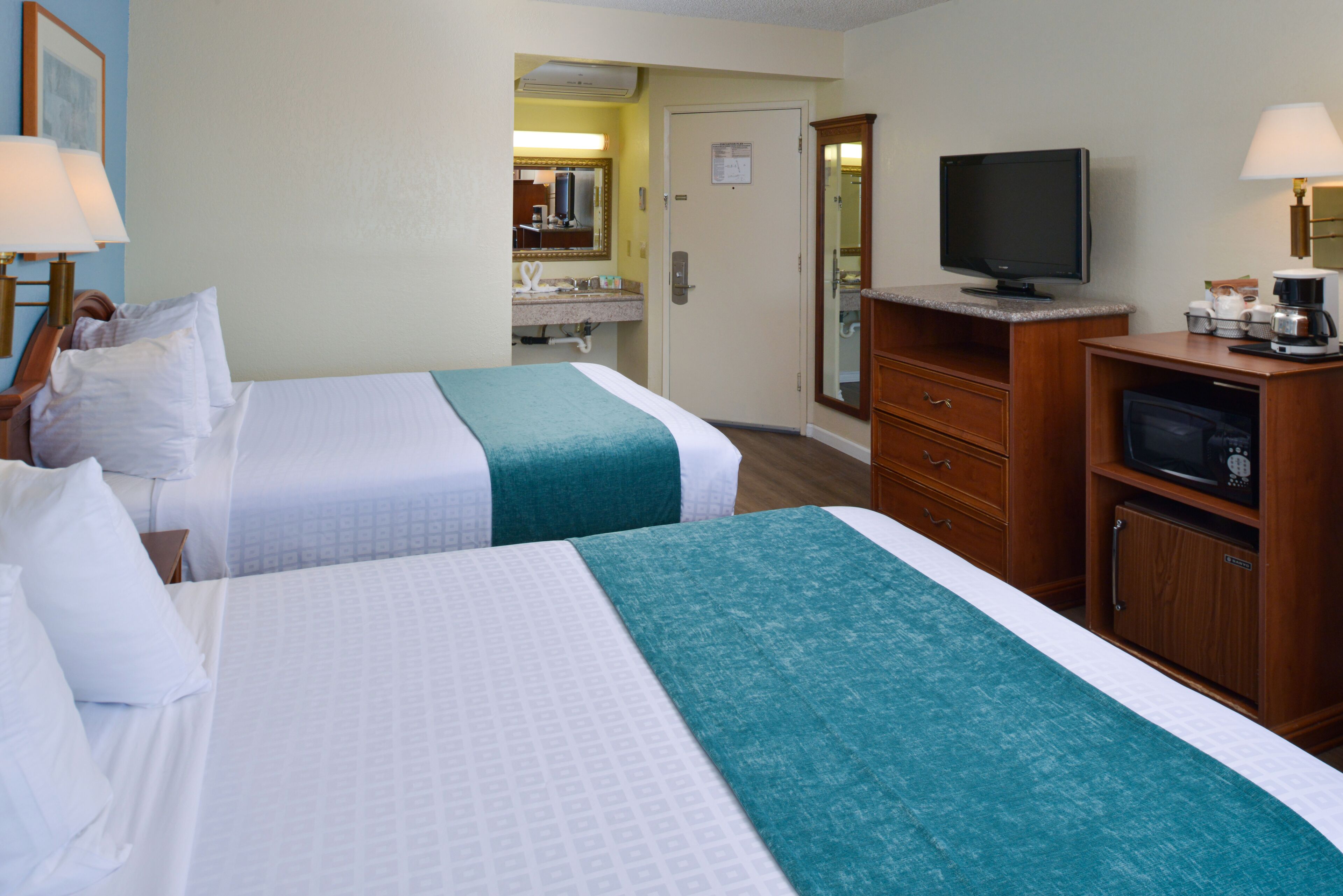 A hotel room with two beds, a television, a desk with a computer, and a bathroom visible through an open door.