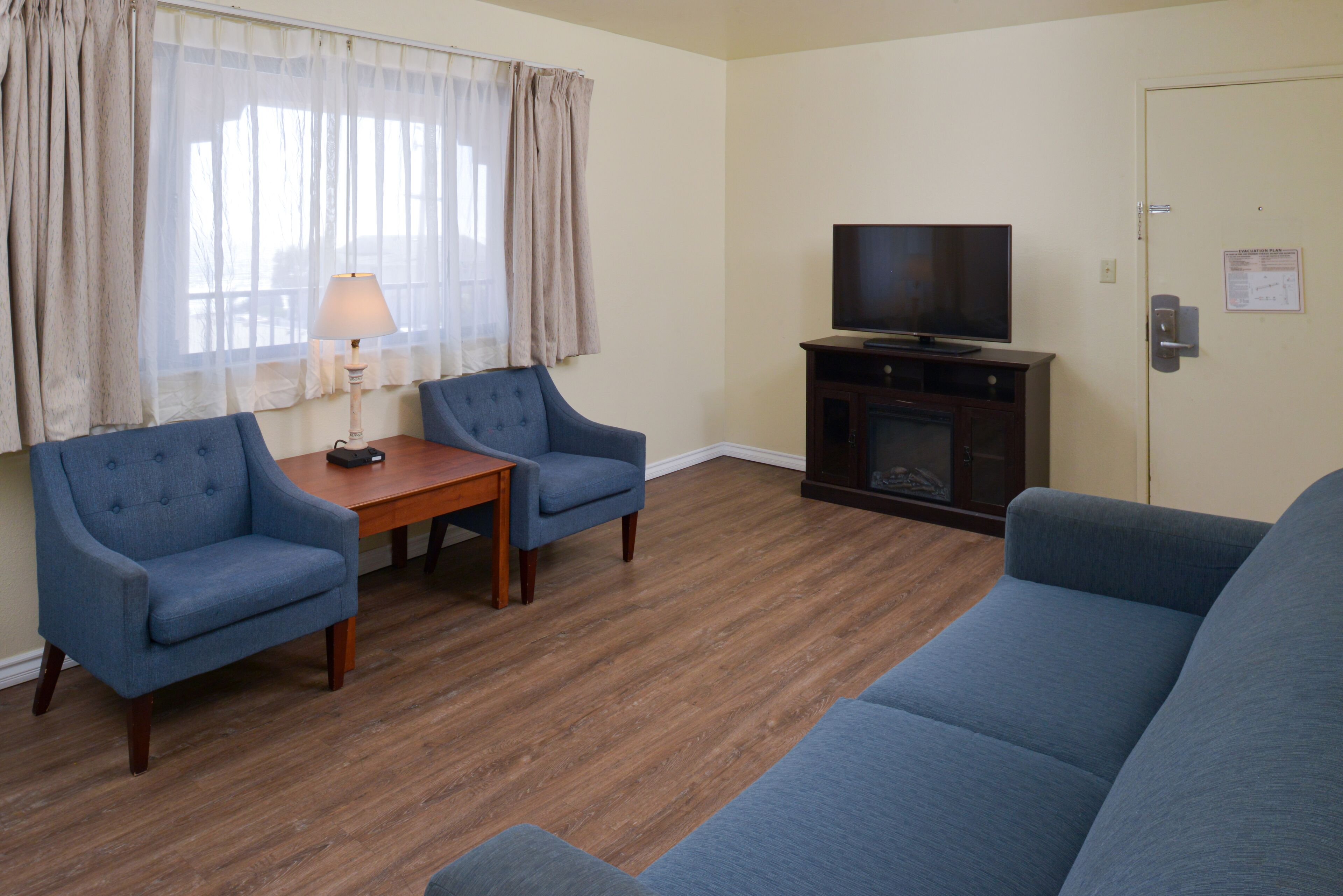 A living room with a wooden floor, a blue sofa, two blue armchairs, a small table with a lamp, and a television on a stand.