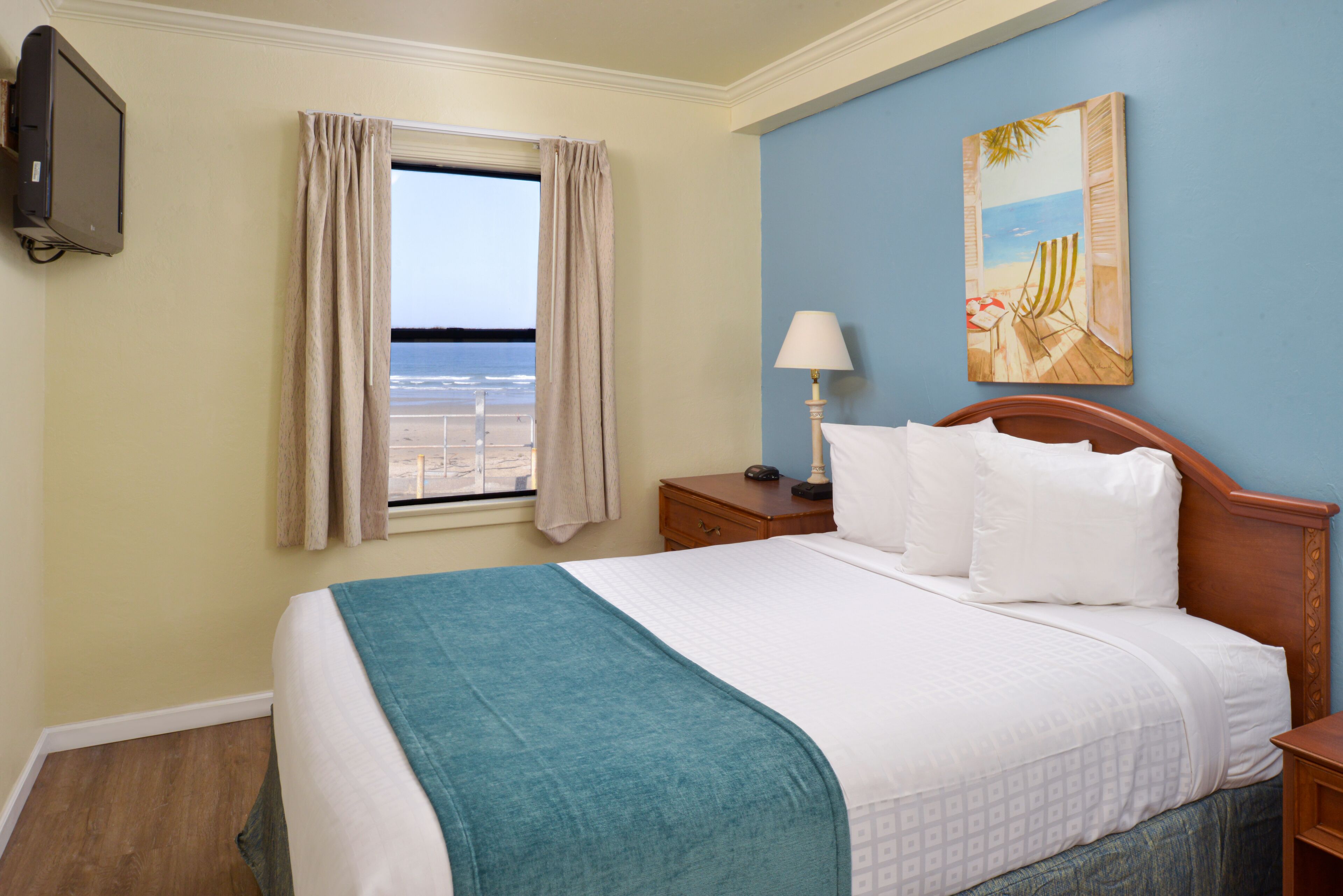 A hotel room with a bed, a nightstand with a lamp, a painting of a beach scene, and a window with curtains.