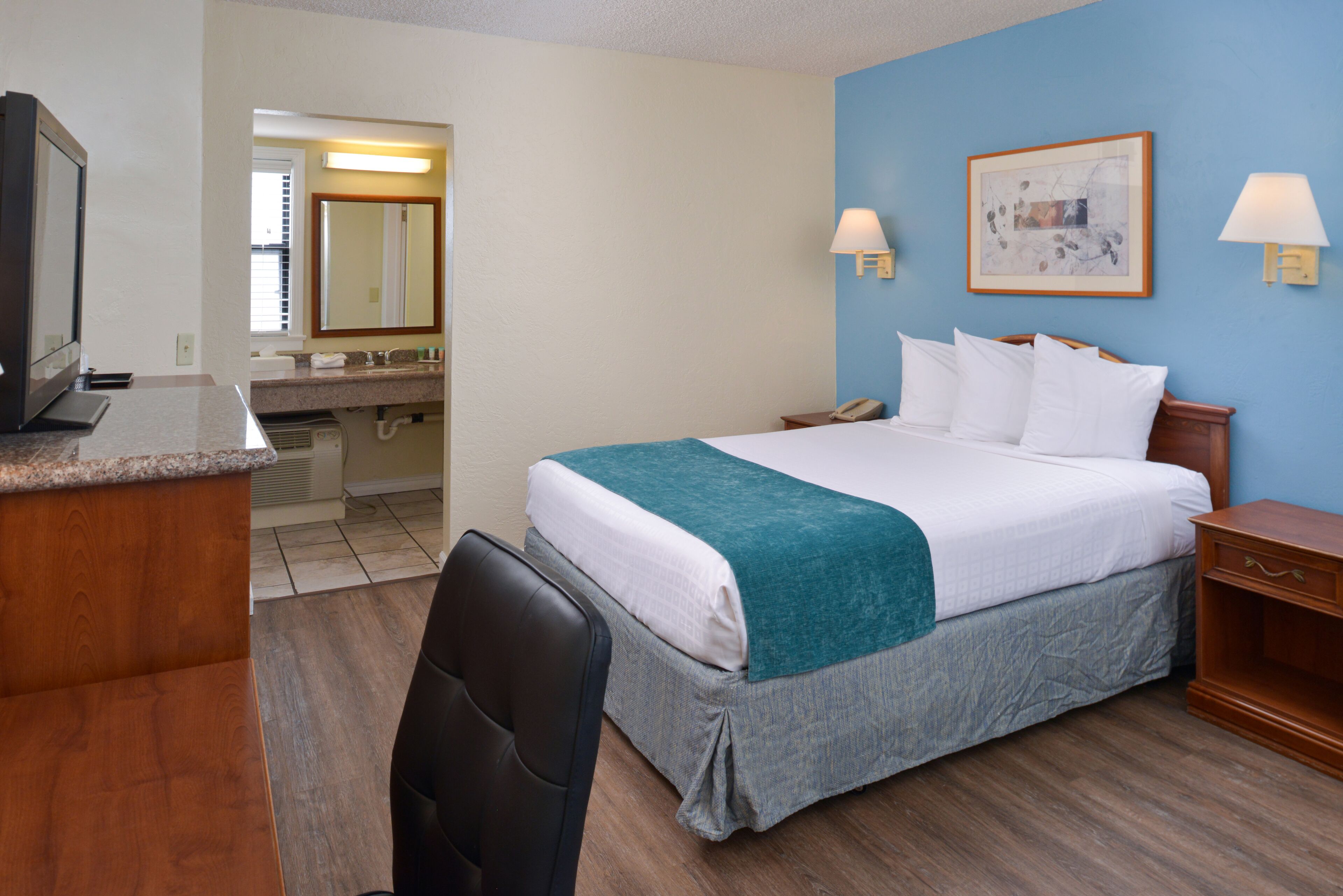 Quarto de hotel com cama, escrivaninha com computador, cadeira, banheiro com pia e espelho, e duas luminárias fixadas na parede.