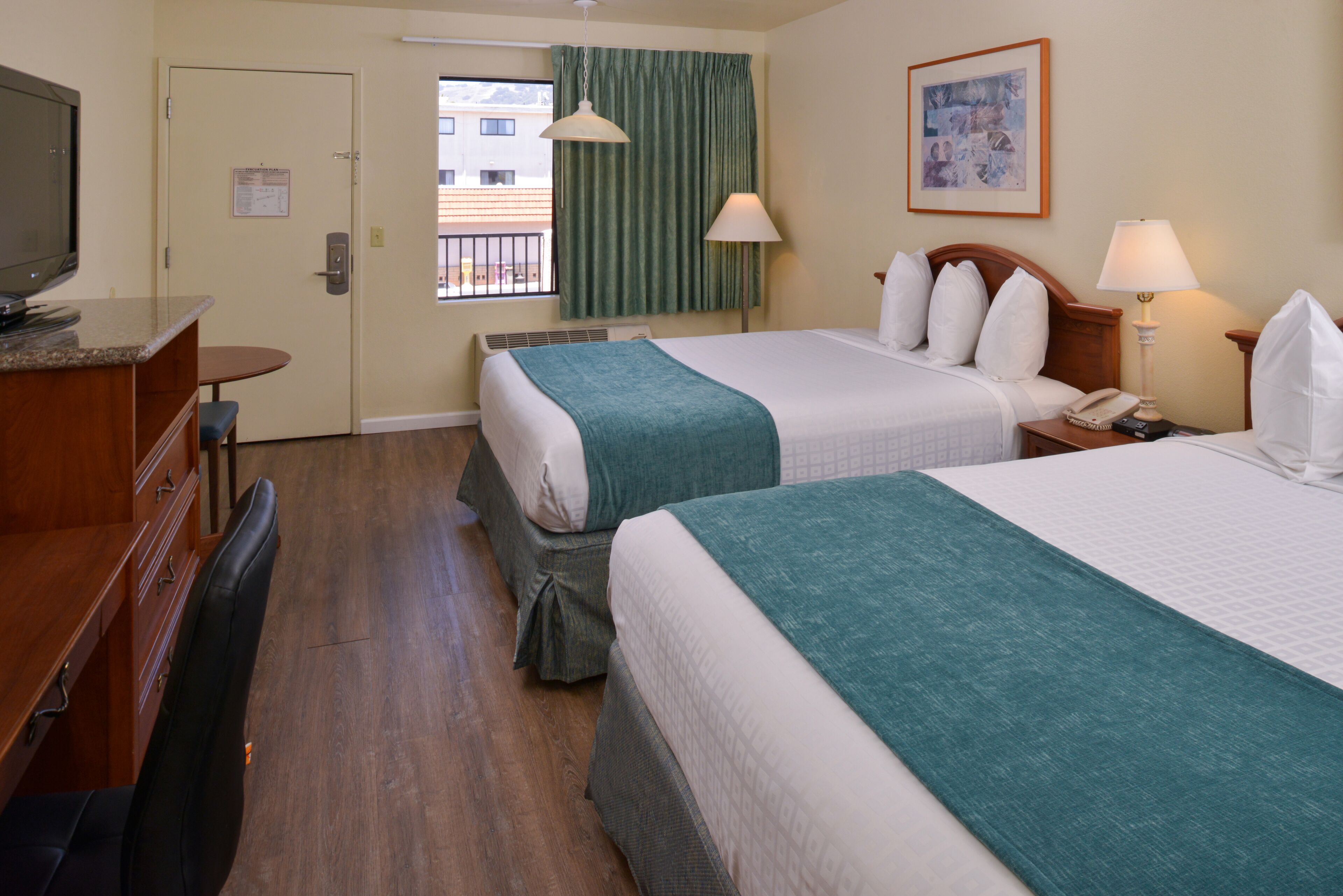 A hotel room with two beds, a desk with a television, a window with curtains, and a painting on the wall.