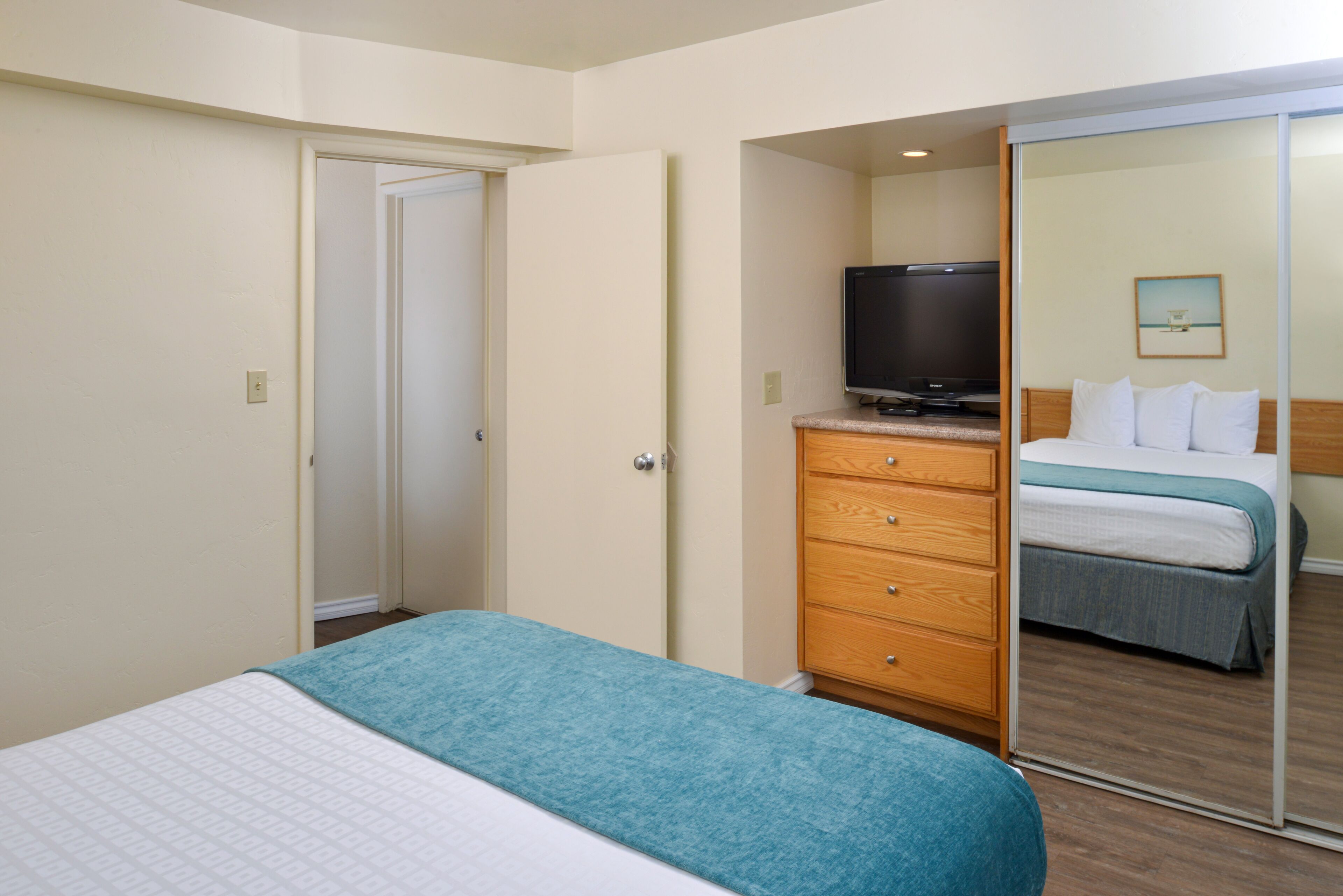 A hotel room with a bed, a television on a dresser, a mirror, and a door leading to another room.