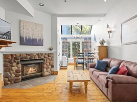 Una sala de estar con chimenea de piedra, piso de madera, un sofá marrón y un comedor con mesa y sillas.
