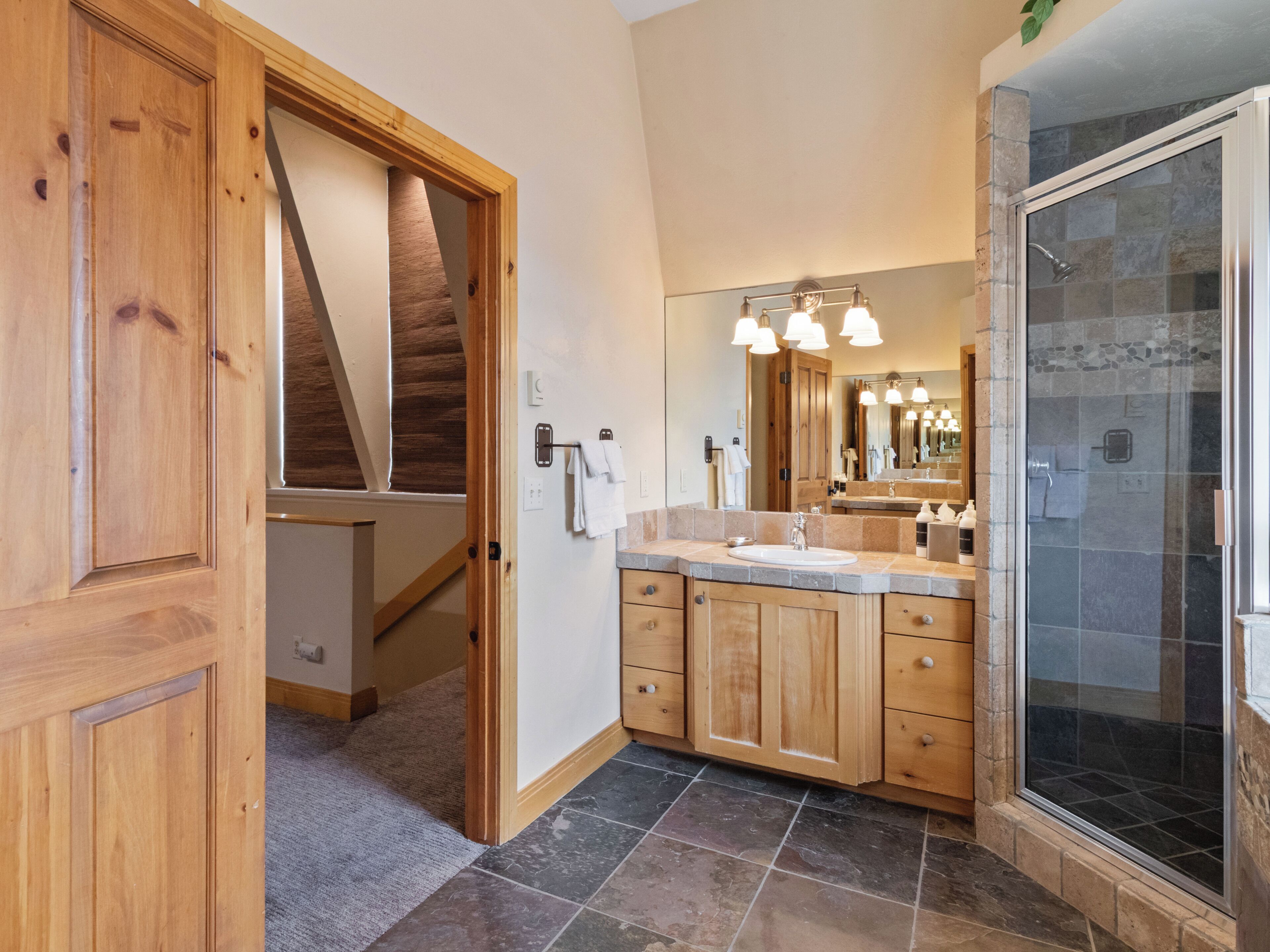 A bathroom with wooden cabinets, a large mirror, a shower with glass doors, and a visible staircase.