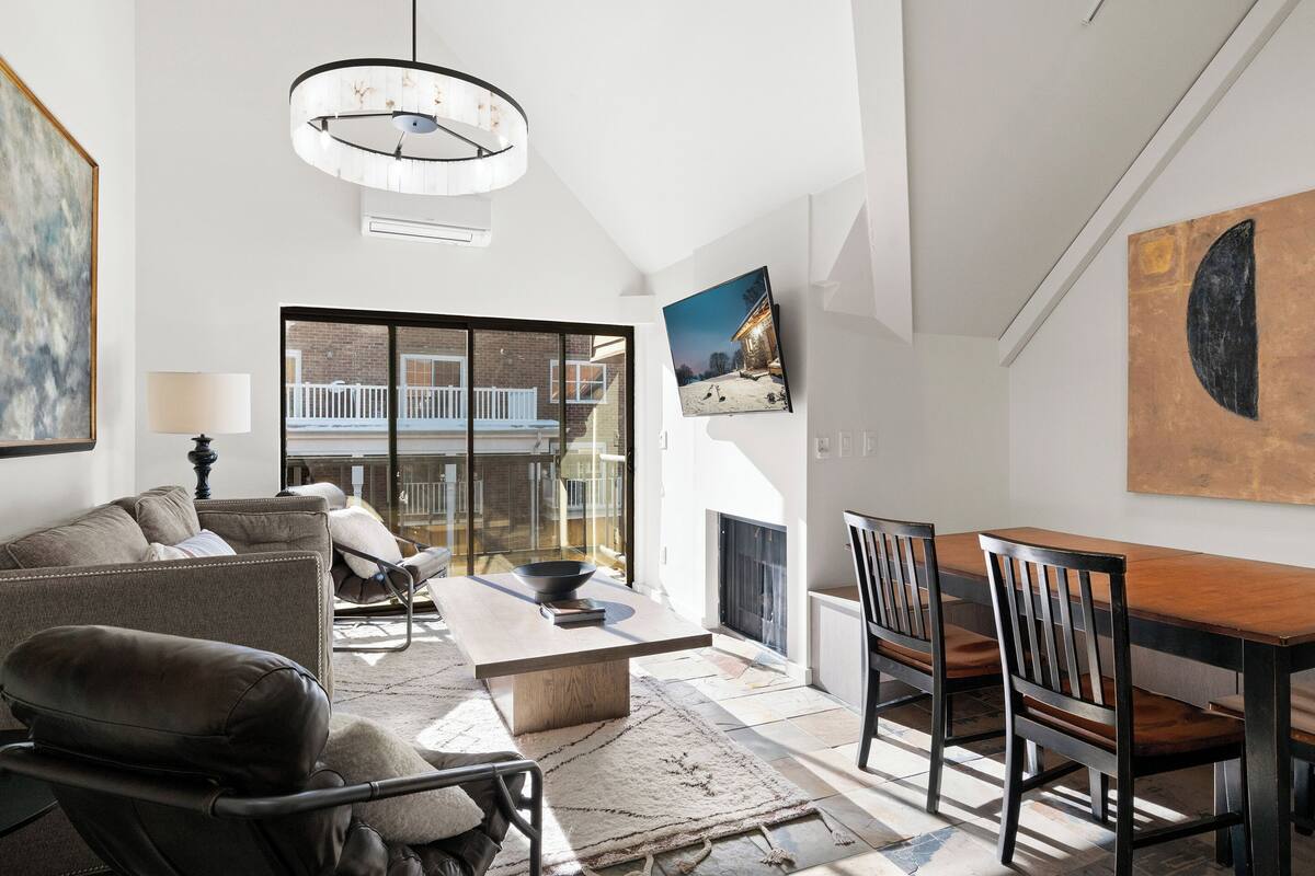 Un salon moderne avec un canapé, une table basse et une table à manger. On y trouve une cheminée, une grande fenêtre et un luminaire au plafond.