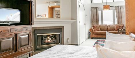 A bedroom with a large bed, a TV, a fireplace, a sofa, and a window with curtains.