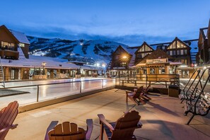 A snow-covered outdoor seating area with wooden chairs and tables, a building with a sign for registration, and a mountain backdrop.