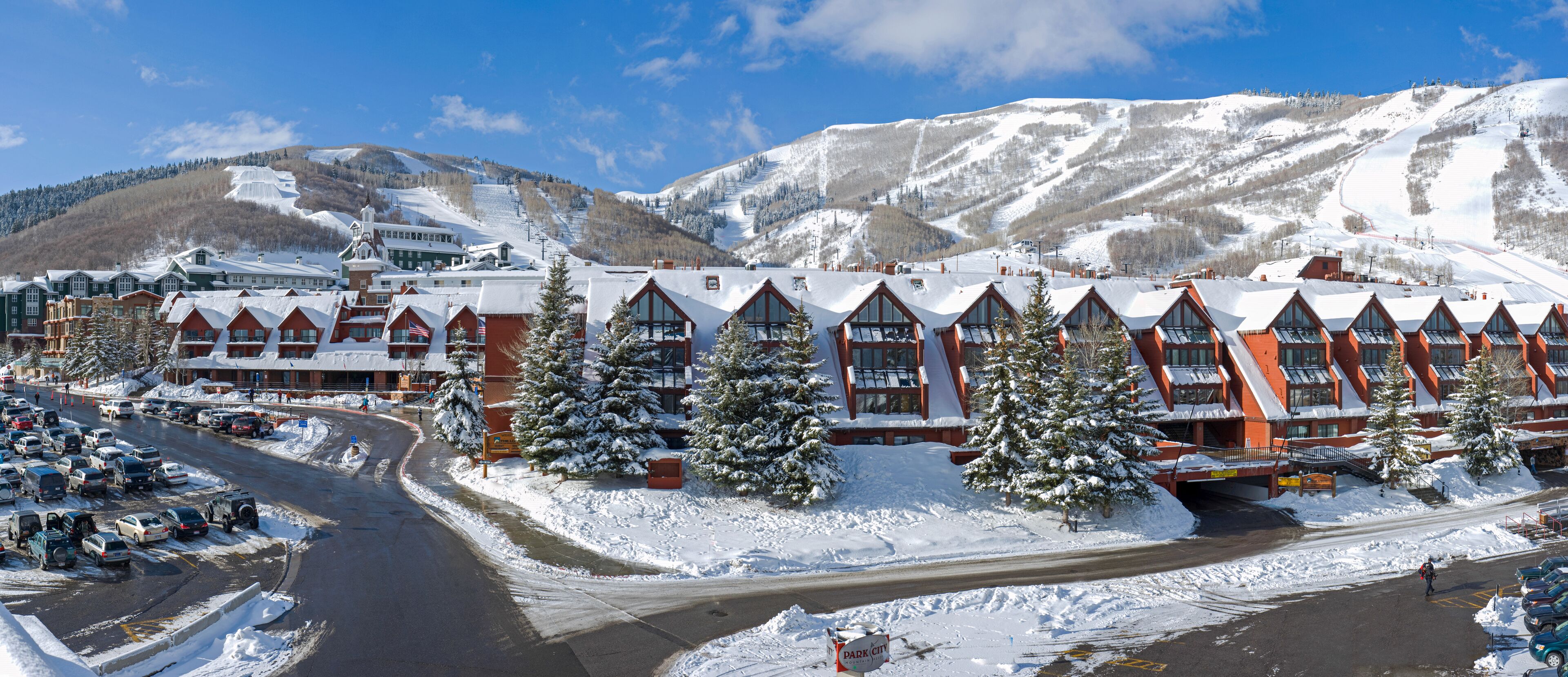 A hotel with a ski slope in the background, a parking lot, and snow-covered trees.