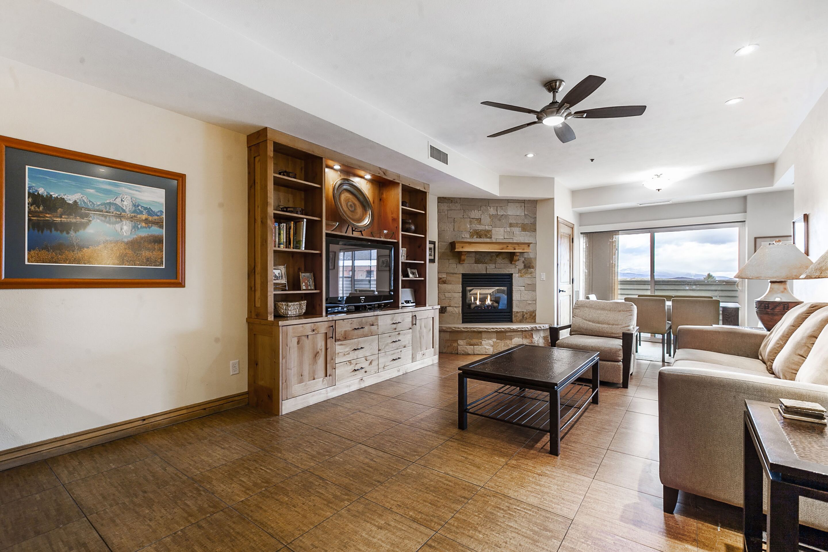 A spacious living room with a fireplace, a ceiling fan, and a large window offering a view.