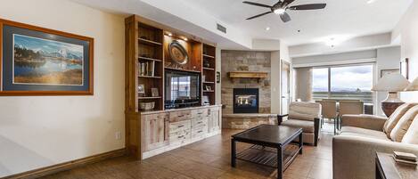 A spacious living room with a fireplace, a ceiling fan, and a large window offering a view.