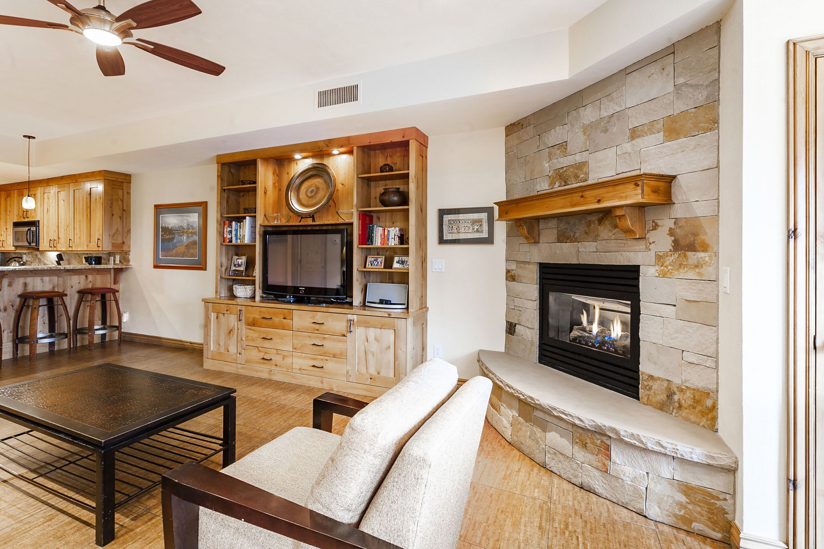 A living room with a stone fireplace, wooden entertainment center, and a ceiling fan.